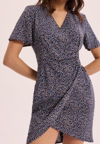 Femme portant une robe portefeuille bleu marine à manches courtes avec un motif à petits pois multicolores, mains reposant sur les hanches sur un fond neutre.