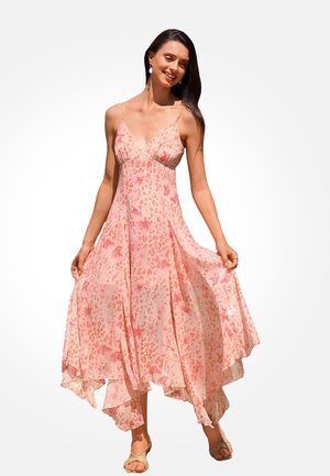 Vrouw glimlachend, draagt een soepel vallende roze jurk met bloemen- en luipaardprint, dunne bandjes en beige sandalen op een witte achtergrond.