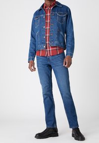 Mann in blauer Jeansjacke und Jeans mit rohem Karoshemd und schwarzen Stiefeln, der vor einem einfarbigen Hintergrund steht.