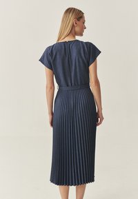 Dunkelblaues Denim-Kleid mit kurzen Ärmeln, das einen plissierten Rock und einen Taillengürtel aufweist. Der Rücken hat einen kleinen Knopfverschluss.