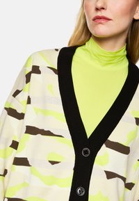 Strickjacke mit schwarzem Rippenkragen, ausgestattet mit einem Camouflage-Muster in limettengrün, beige und braun. Zwei sichtbare Knöpfe zum Schließen.