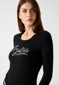 Schwarzes Langarmshirt aus weichem Stoff mit einem silbernen, aufgestickten "Guess"-Logo auf der Brust. Rundhalsausschnitt und figurbetonte Passform.