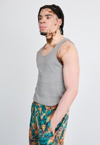 Maiou gri dintr-un material moale, asociat cu pantaloni scurți colorați, cu un imprimeu tropical în nuanțe de verde, portocaliu și albastru. Modelul stă cu mâna în buzunar.
