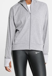 Persoon in een lichtgrijs hoodie met rits en duimopening over een wit shirt en zwarte Nike leggings met wit logo, staand tegen een witte achtergrond.