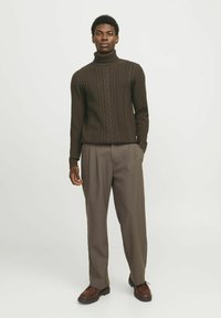 Maglione a collo alto in maglia a treccia marrone con polsini a coste, abbinato a pantaloni beige larghi e scarpe marroni, in piedi davanti a uno sfondo chiaro.