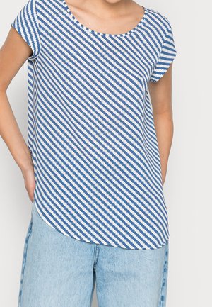 Femme portant un haut à manches courtes à rayures diagonales bleues et blanches associé à un jean denim bleu clair, mains dans les poches.