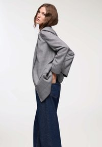 Giovane donna con capelli castani di lunghezza media, che indossa un blazer grigio a quadri e jeans larghi blu scuro, in posa con una mano nella tasca posteriore.