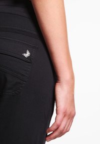 Svarta jeans med sidficka, prydda med en silverfärgad fjäril som accent. Tyget har en slät yta och en åtsittande design.