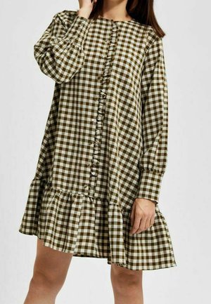 Femme portant une robe longue à manches longues en vichy marron et blanc avec un devant à boutons et un ourlet volanté, posant avec une main près du visage.