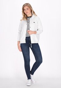 Felpa zip grigia chiara con logo di un ombrello nero, abbinata a una maglietta a righe bianco e nera, jeans scuri e sneaker bianche.