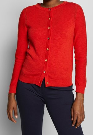 Woman wearing a fitted red buttoned cardigan with ribbed cuffs and hem, paired with dark navy pants.