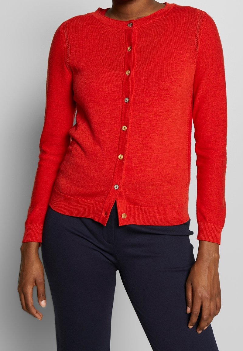 Femme portant un cardigan rouge ajusté à boutons avec des poignets et un bas côtelés, associé à un pantalon bleu marine foncé.