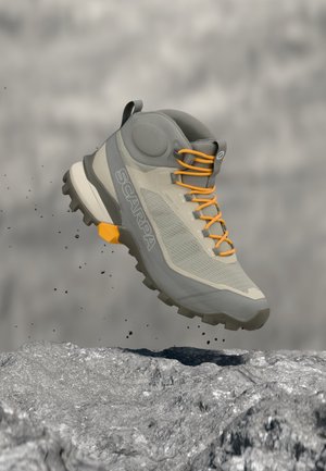 Šedá turistická obuv s oranžovými šnúrkami vo vzduchu nad skalnatým terénom, so zodolnenou podrážkou a nápisom "SCARPA" na boku.