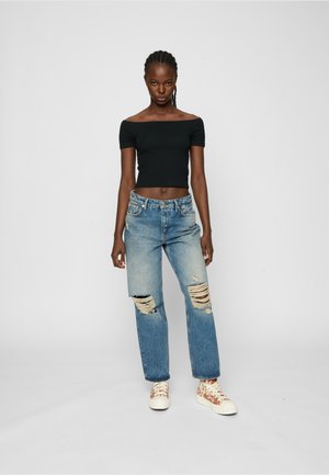 Schwarzes schulterfreies Oberteil, zerrissene hellblaue Jeans und beigen Sneakers mit Muster. Modell posiert vor einem schlichten weißen Hintergrund.