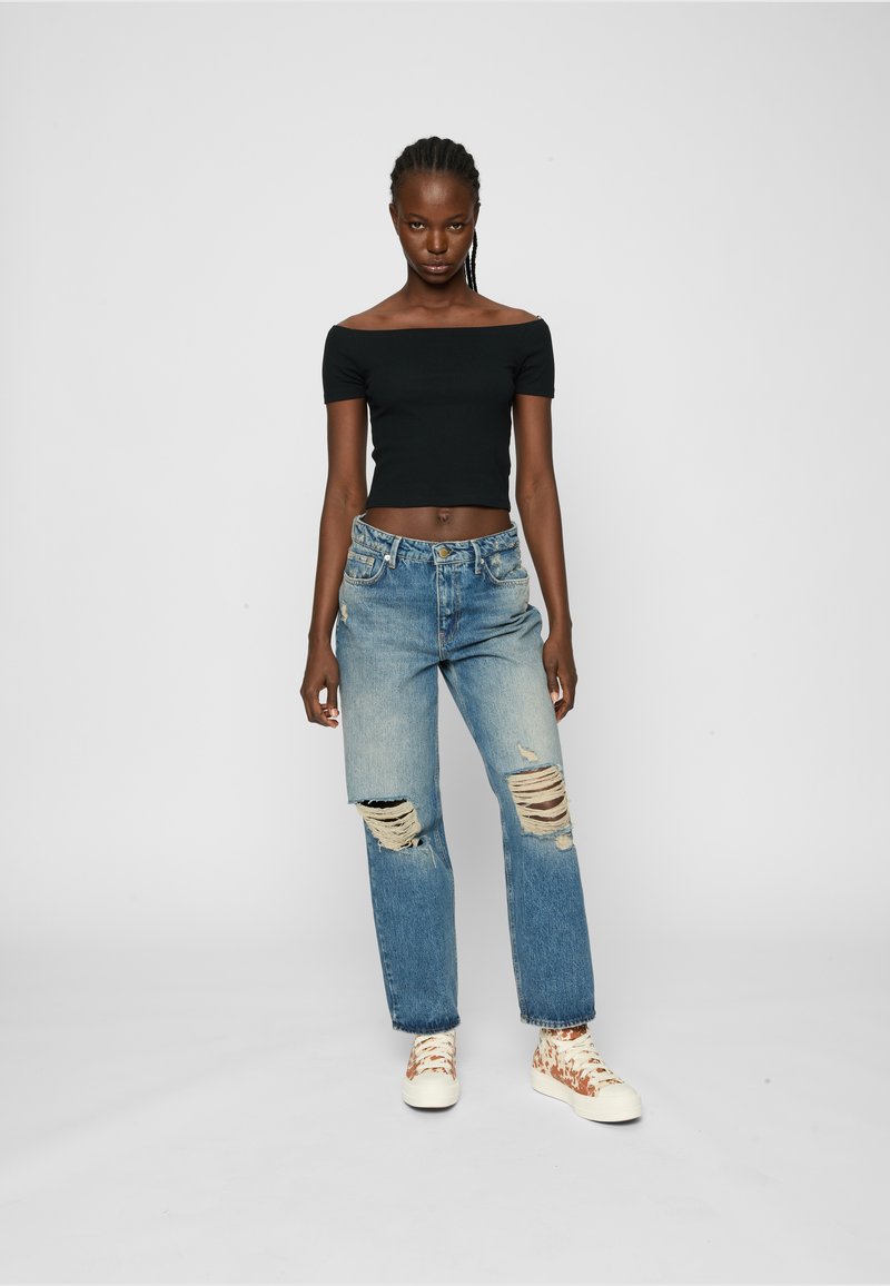 Top negru, off-the-shoulder, blugi deschiși cu rupturi și adidași bej cu model. Modelul pozează pe un fundal alb simplu.