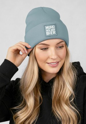 Hellblauer, gerippter Beanie mit einem quadratischen weißen Aufnäher mit Text. Wird leicht schräg getragen, mit glatter Textur und lässiger Passform.