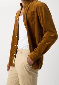 Hombre con camisa de pana mostaza de botones sobre una camiseta blanca sin mangas y pantalones beige, con la mano en el bolsillo, frente a un fondo liso.