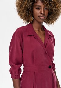 Kobieta o kręconych włosach, ubrana w głęboko czerwoną sukienkę kopertową z rękawami 3/4 i czarnym guzikiem, stojąca na jednolitym białym tle.