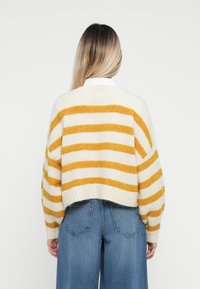 Crème en mosterd gestreepte gebreide trui met een cropped ontwerp, brede mouwen en geribde zoom. Gecombineerd met high-waisted blauwe jeans.