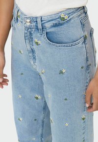 Lichtblauwe denim jeans met geborduurde witte madeliefjes en groene stelen, hoge taille en traditionele vijf-pocket stijl.
