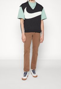 Homme portant un pull sans manches noir avec un grand swoosh blanc de Nike sur un t-shirt vert menthe, un pantalon marron et des baskets montantes blanches.