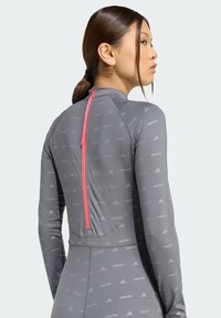 Sieviete ar brūniem matiem, savienotiem zirgaste, valkā pelēku sporta krekli ar atkārtotiem "MOON BOOT" un Adidas logotipiem, kā arī vertikālu sarkanu rāvējslēdzēju aizmugurē.