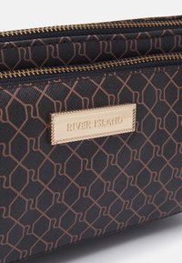 Svart och brun mönstrad dragkedjefodral med en guld metallskylt graverad med "RIVER ISLAND" på framsidan.
