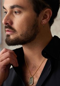 Pendentif en forme de plaque d'identité en or avec un design complexe sur une chaîne en or. Porté avec une chemise noire, accompagné d'une petite boucle d'oreille en or en forme de croix.