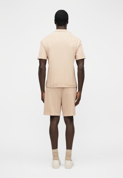 Homme vu de dos portant une chemise à manches courtes texturée beige, un short assorti, des chaussettes beiges et des baskets blanches sur un fond uni.