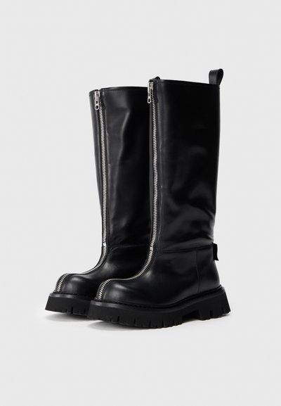 Bottes en cuir noires jusqu'aux genoux avec des semelles épaisses et des fermetures éclair verticales en argent allant du bout à la tige sur un fond uni.