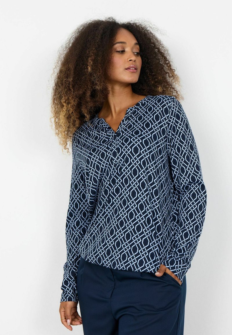 Tmavomodrá blúzka s geometrickým bielym vzorom, s výstrihom do V a dlhými rukávmi. Vyrobená z ľahkej, hladkej látky.