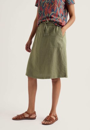Personne portant une jupe vert olive longueur genoux avec des poches avant, un haut coloré à motifs, et des sandales plates marron avec des lanières.