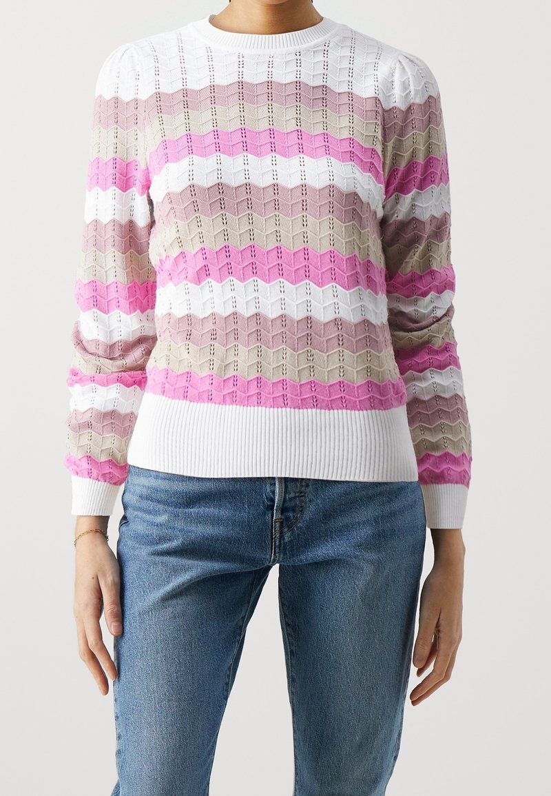 Femme portant un pull en maille à rayures horizontales ondulées roses, blanches, olives et mauves, associé à un jean bleu.