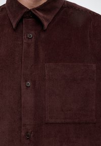 Gros plan d'une chemise en velours côtelé marron foncé avec un col, trois boutons noirs et une poche poitrine carrée sur le côté droit.