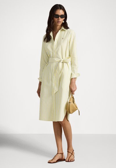 Kvinne iført gul og hvit stripete skjortedrakt med belte, svarte solbriller, brune sandaler, holder en brun trekantet veske mot hvit bakgrunn.