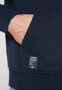 Sudadera con capucha de color azul marino, con puños acanalados, bolsillo tipo canguro y una etiqueta de tela que muestra "CAMP DAVID" y los detalles del producto.