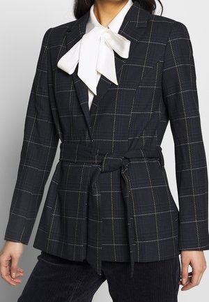 Kobieta ubrana w ciemną kraciastą marynarkę z wiązaniem w talii na białą bluzkę z dużą kokardą przy kołnierzu, połączona z ciemnymi prążkowanymi spodniami.