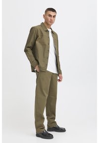 Olivengrøn bomuldsjakke med knaplukning, kombineret med matchende bukser. Modellen står med hænderne i lommerne og har en hvid T-shirt under.