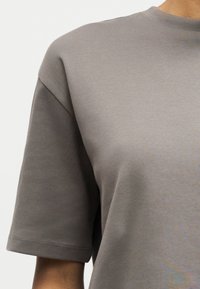 Κοντινό πλάνο ενός ατόμου που φοράει φαρδιά, κοντομάνικη μπλούζα σε χρώμα ταουπέ με στρογγυλή λαιμόκοψη.