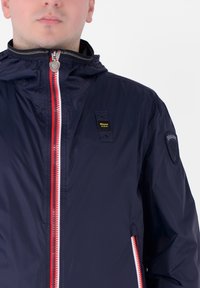 Giacca leggera blu navy con una zip rossa e bianca, cappuccio e patch con logo sul lato sinistro del petto. Presenta tasche laterali con zip.