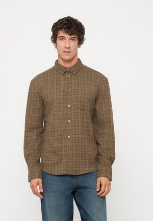 Homme portant une chemise à carreaux marron à boutons avec manches longues et un jean bleu, debout devant un fond blanc uni.