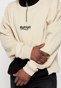 Sweatshirt beige à fermeture éclair avec accents noirs. Comprend un col rond, des poignets côtelés et le logo « Multiply » en noir sur le devant.