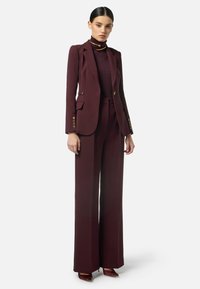 Completo su misura borgogna con giacca aderente, pantaloni a gamba larga e dolcevita texturizzato. Accenti di bottoni dorati e design lineare.