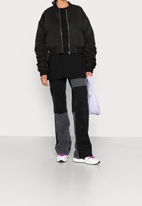 Giacca bomber nera corta con zip, abbinata a pantaloni patchwork neri e grigi. La modella tiene una borsa di colore viola chiaro. Scarpe bianche con accenti viola.