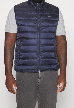 Mand iført navyblå quiltet ærmeløs vest over grå kortærmet skjorte med grå jeans, stående mod en ren hvid baggrund.