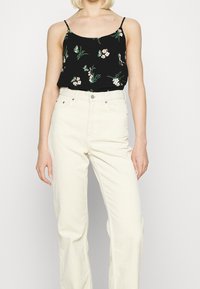 Camisole noire à motif floral avec fines bretelles, associée à un pantalon en velours côtelé beige taille haute, avec poches avant et fermeture à boutons.
