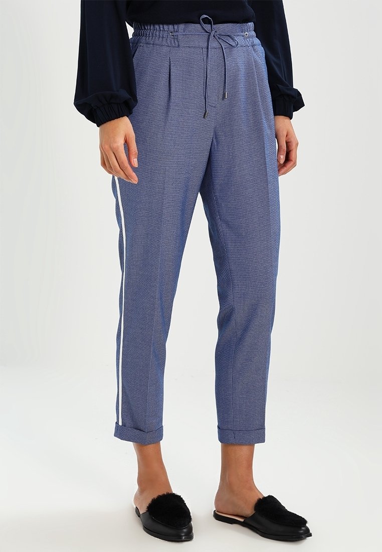 Pantalon bleu fuselé avec bandes blanches sur les côtés, taille élastique avec cordon, porté avec des chaussures noires à enfiler avec détail en fourrure.