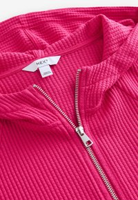 Sweat à capuche fuchsia à texture gaufrée avec une fermeture éclair argentée et un grand col. L'étiquette indique la marque « NEXT » et « Fabriqué au Cambodge ».