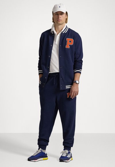 Navy tracksuit with white striped accents, featuring an orange "P" on the jacket and pants. Worn with a white cap and blue sneakers.
