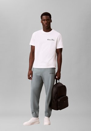 Mężczyzna w białej koszulce Calvin Klein i szarych spodniach dresowych, trzyma czarny plecak w prawej ręce, stoi na szarym tle.